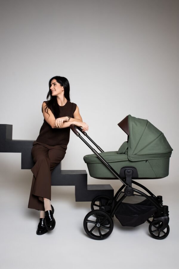 Žalias Tutis Gio+ kūdikių vežimėlis su lopšiu ir juodu rėmu, šalia sėdinti moteris