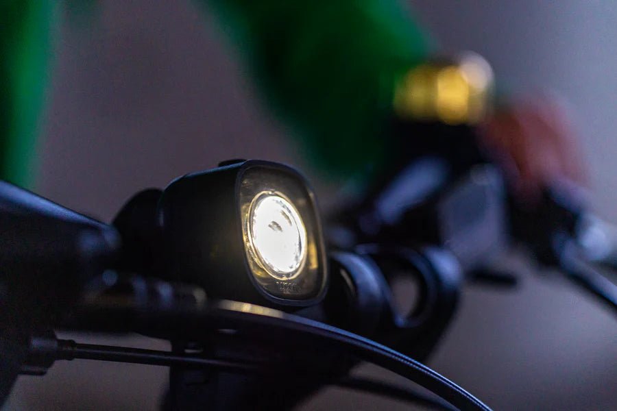 WOOM BEAM velosipēda lukturis melnā krāsā ar ieslēgtu priekšējo gaismu uz velosipēda stūres