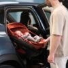 Oranžs Maxi-Cosi zīdaiņu autokrēsliņš automašīnā ar bērnu un vīrieti pie atvērtām durvīm